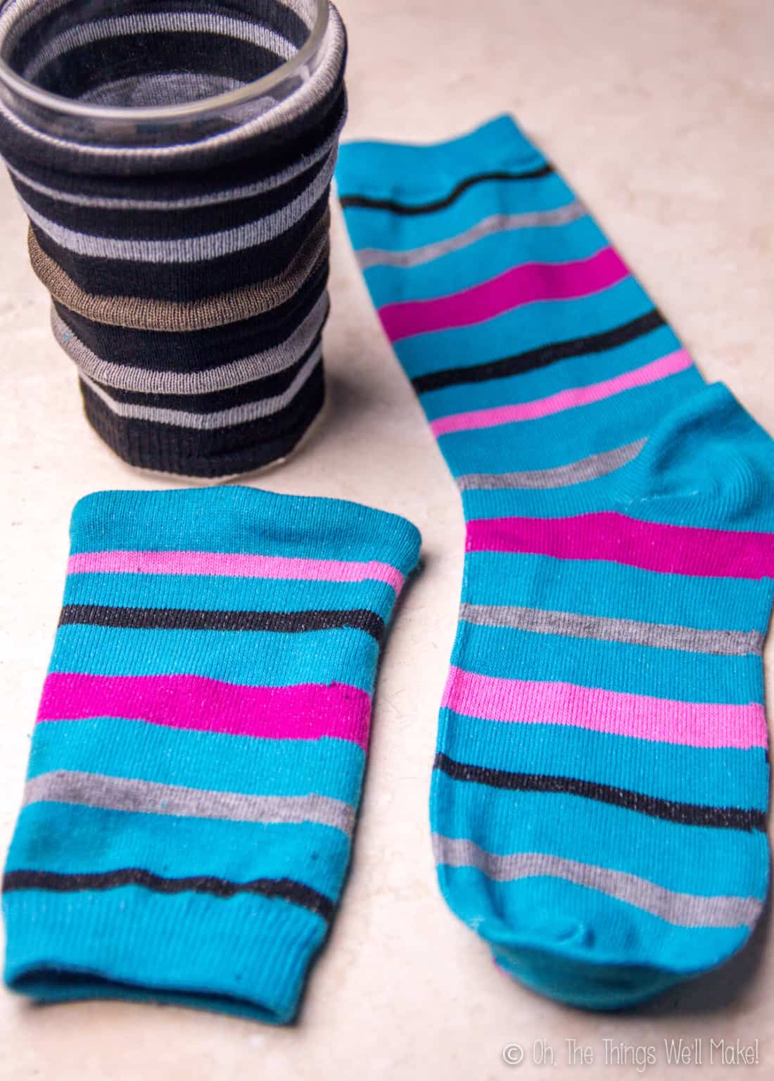 The finished black striped coffee cozy slipped over a glass with a finished blue striped coffee cozy laid down next to a striped blue sock.