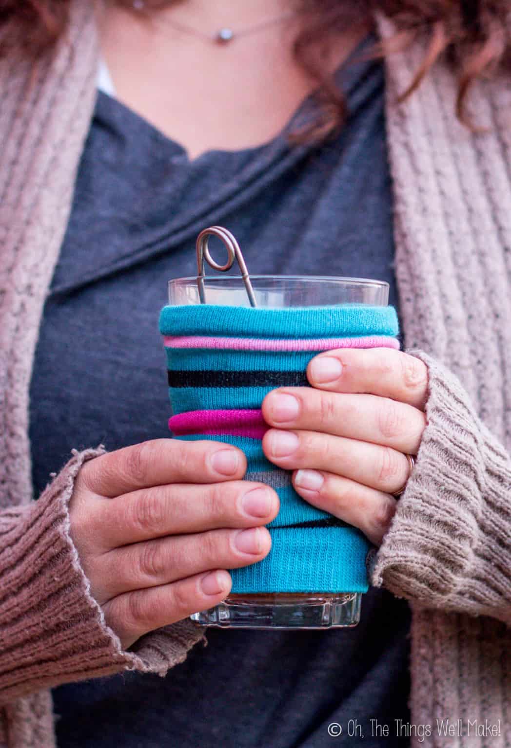 Close up of female hands holding a steamy cup of tea with a homemade striped coffee cozy over the cup.