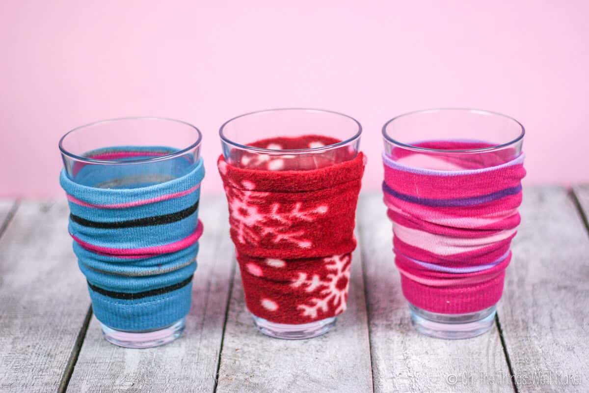 Three coffee cozies on glasses standing on a wood surface. The left one is a blue striped cozy made out of a sock, the middle one is a red polar fleece fabric cozy, and the right one is pink striped cozy made out of a sock.