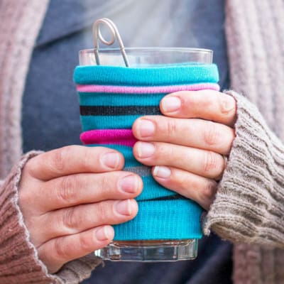 A closeup of a woman's hands holding a hot glass of coffee covered in a bright striped blue and purple cozy made out of a sock.