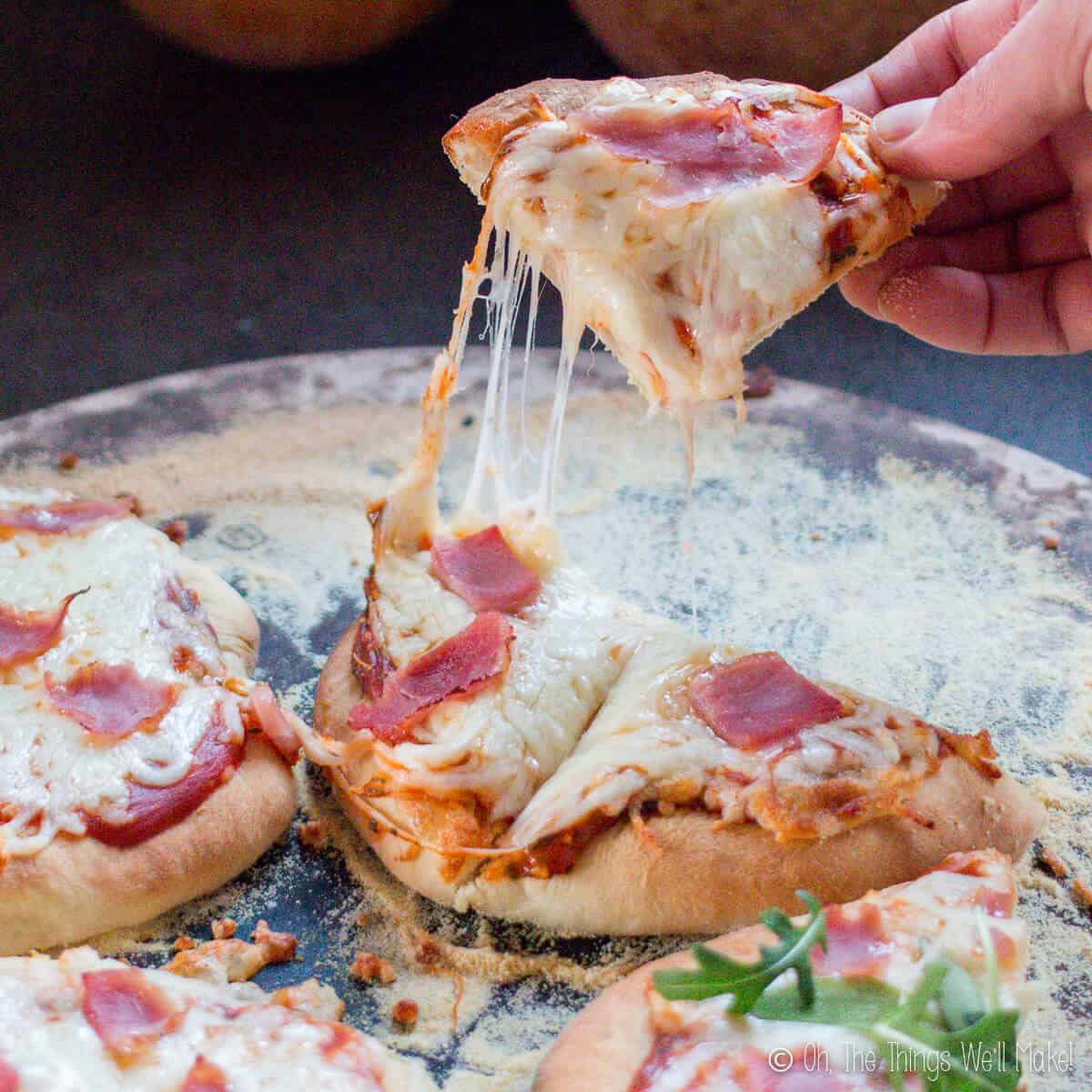 A hand lifting up a slice of mini pizzas with strings of cheese hanging down.