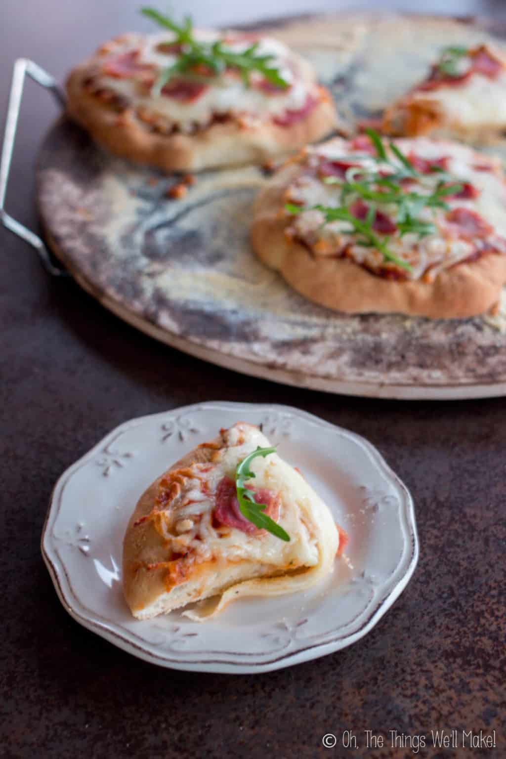 A slice of pizza on a white plate below several individual mini sized pizzas on a circular wood tray.