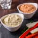 2 bowls of zucchini hummus, one cream colored and the other orange colored because it's made with roasted red peppers. They're surrounded by cut raw veggies.