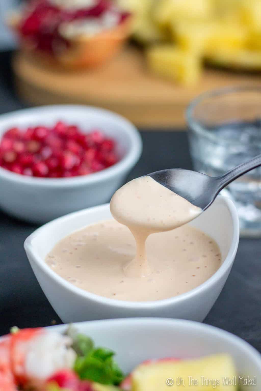 Close up of a spoon full of salsa rosa dripping down into a white bowl full of the same sauce and a bowl of pomegranates in the back.