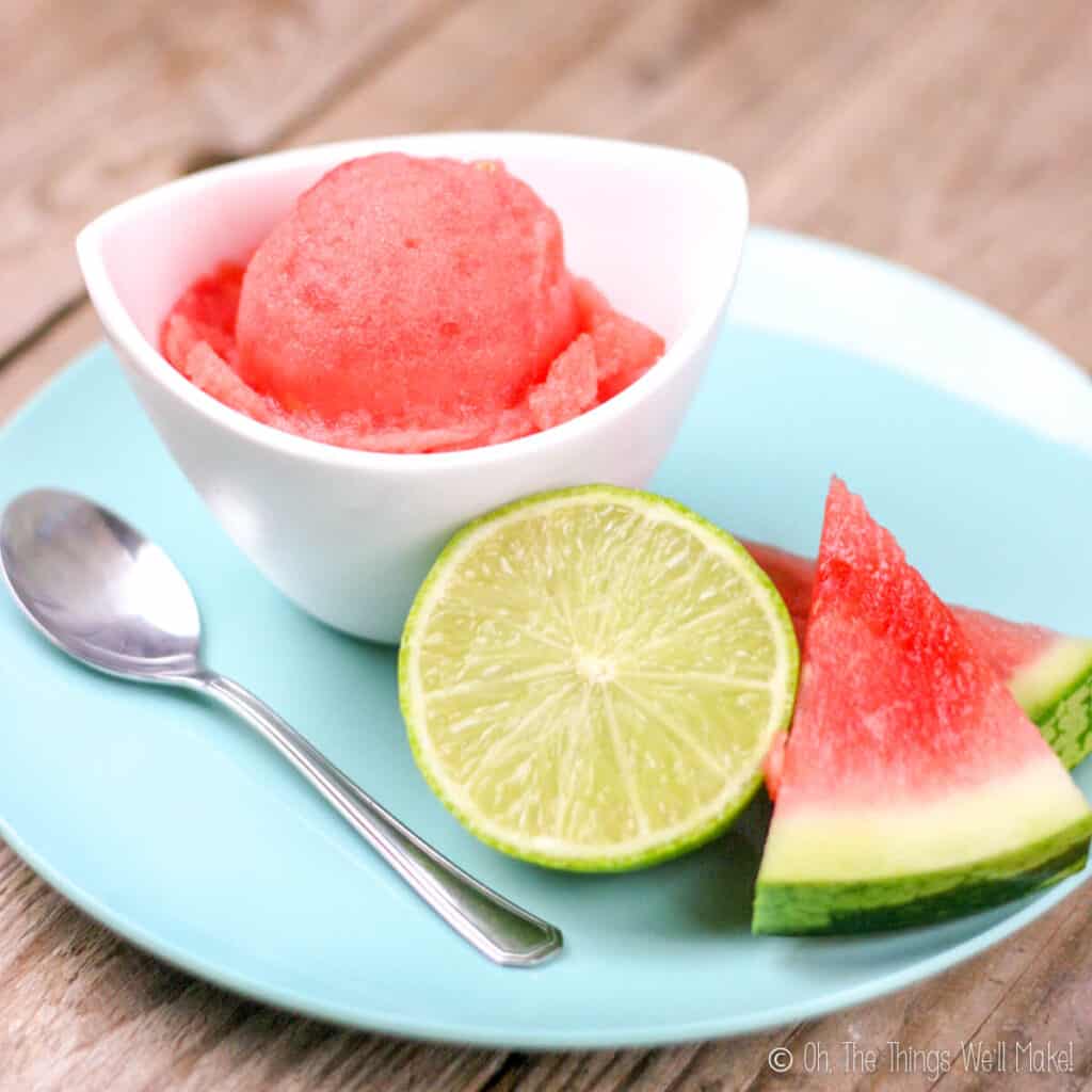 A watermelon sorbet in a white bowl on a plate with some slices of watermelon and half a lime.