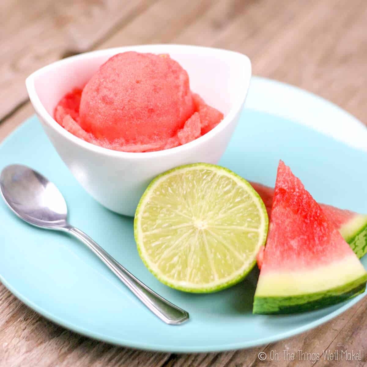 A watermelon sorbet in a white bowl on a plate with some slices of watermelon and half a lime.