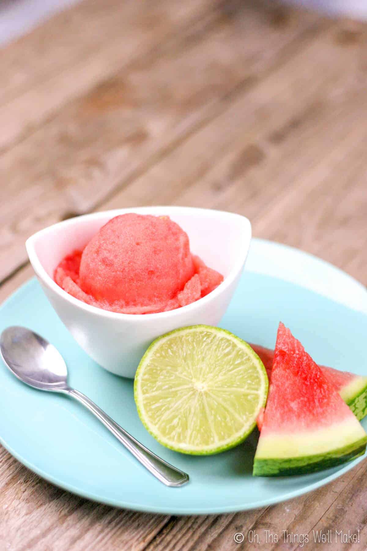 A small bowl of a homemade, easy watermelon sorbet on a plate with a slice of watermelon and half a lime