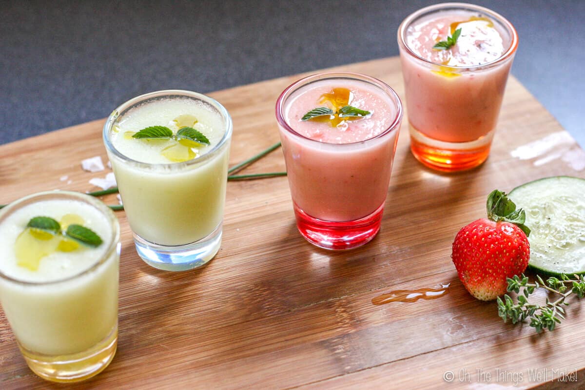 Side view of 4 shot glasses filled with a chilled gazpacho soup. The two on the left are green, the two on the right are pink. All are garnished with mint. 