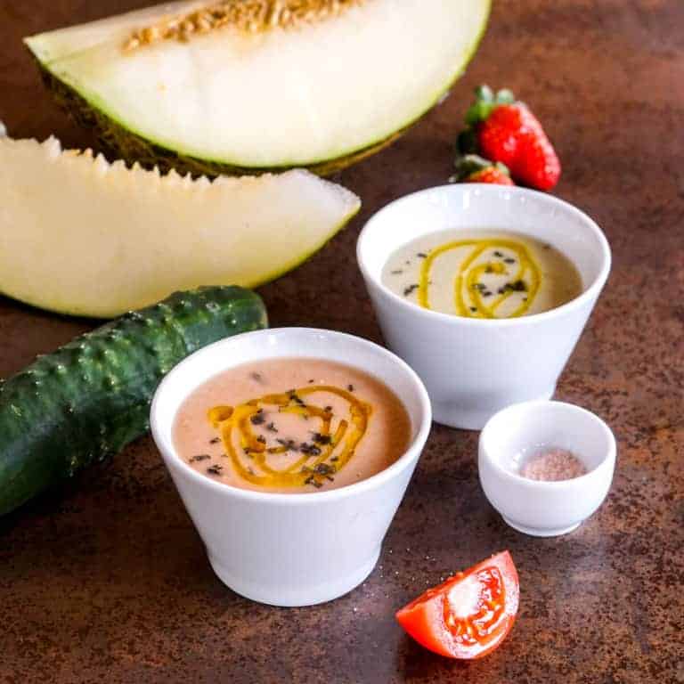 Two bowls of homemade strawberry melon gazpacho soup with a small bowl of pink himalayan salt beside it. Cut up melon, cucumber, some strawberries, and a slice of tomato are scattered around the bowls.