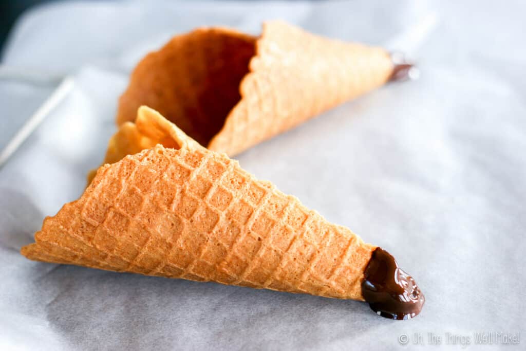 Two homemade waffle cones with the tips dipped in chocolate, resting on a parchment lined tray to cool and harden.