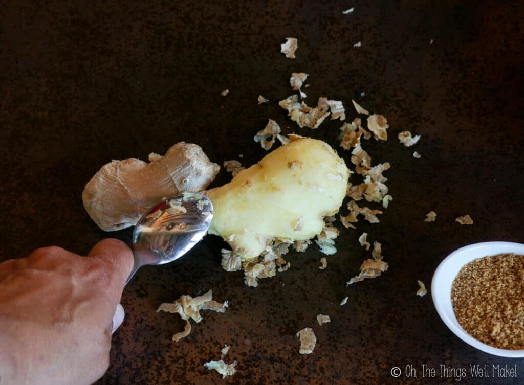 A piece of ginger root whose skin is being peeled with a spoon. Pieces of the skin are scattered around it.