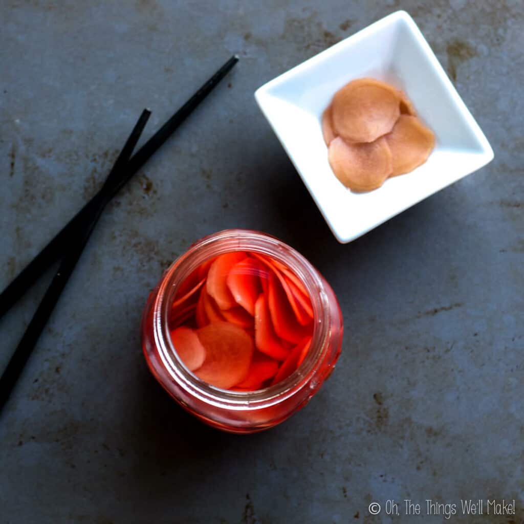overhead view of an open jar of pickled ginger that has been colored pink with beetroot. It's next to a small bowl filled with pickled ginger slices and some chopsticks.