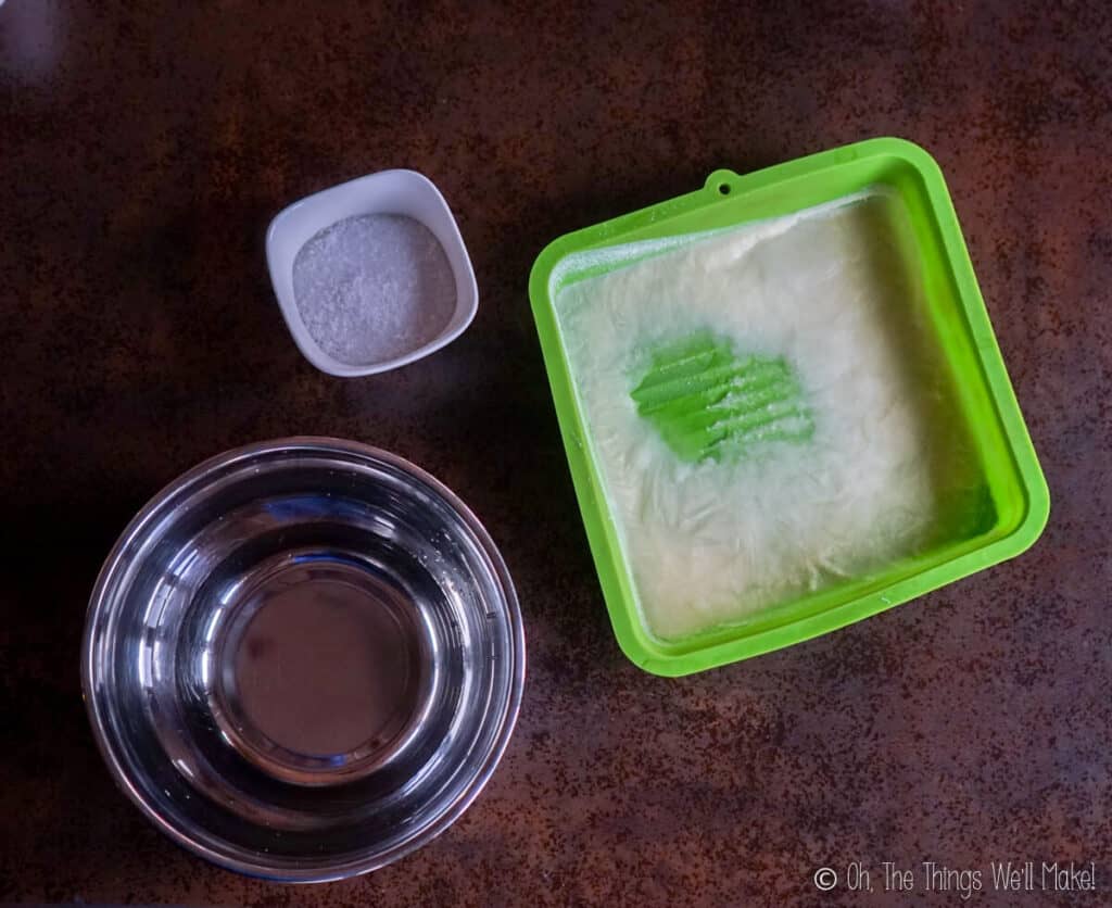 Overhead view of frozen goat milk in a pan, a bowl of lye, and a bowl of water.