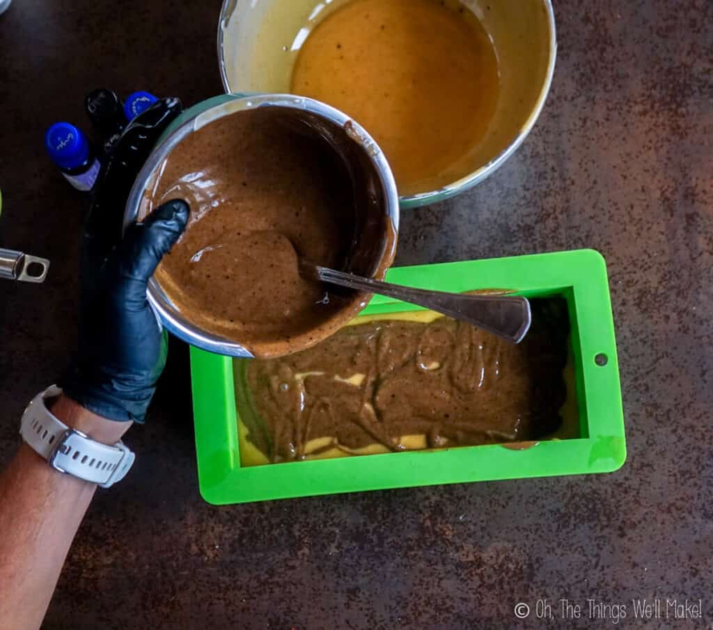 Pouring a dark colored spiced soap mixture into a soap mold over a lighter colored soap mixture
