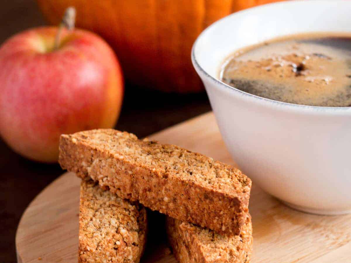 Close-up of three pieces of pumpkin spice biscotti cookies placed beside a cup of coffee on a wooden platter with an apple and pumpkin in the background.