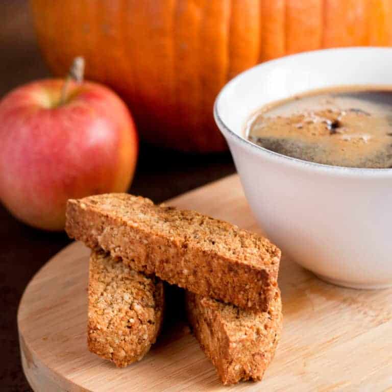 Close-up of three pieces of pumpkin spice biscotti cookies placed beside a cup of coffee on a wooden platter with an apple and pumpkin in the background.