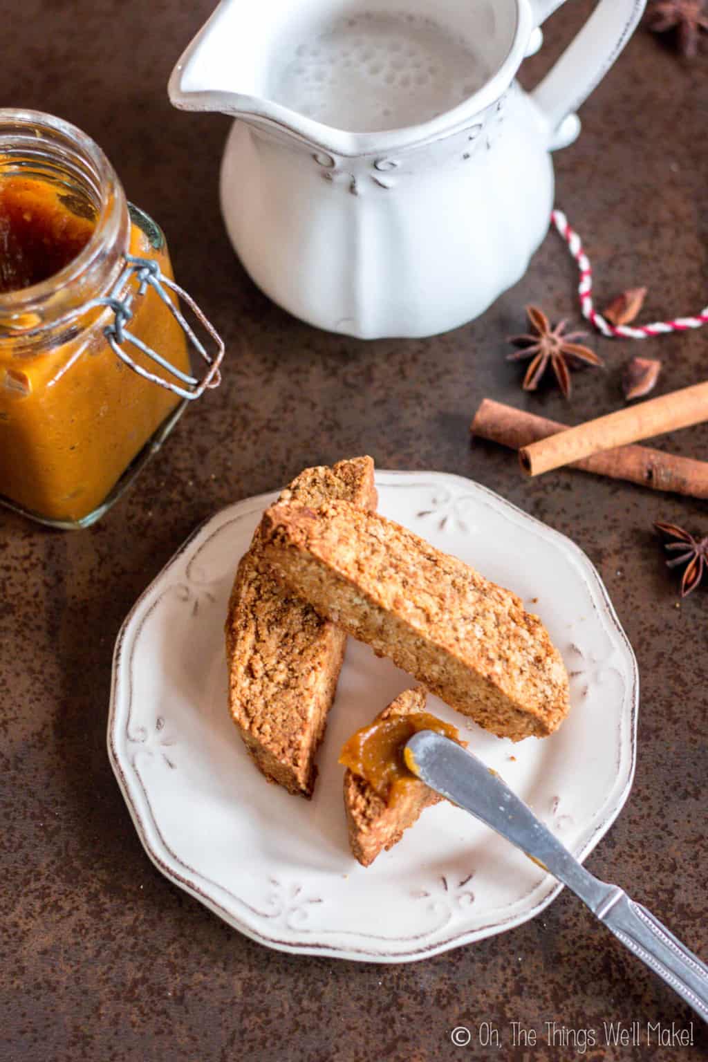 Top view of three pumpkin spice biscotti on a plate with a knife spreading spiced pumpkin butter over one piece. Beside the plate are an open jar of pumpkin butter, a white ceramic jug, and a couple pieces of cinnamon sticks and star anise seeds.