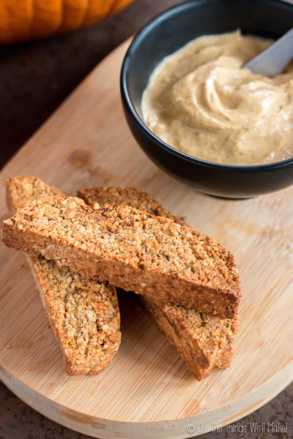 Three pumpkin spice biscotti on a wooden board with a black bowl full of pumpkin butter.