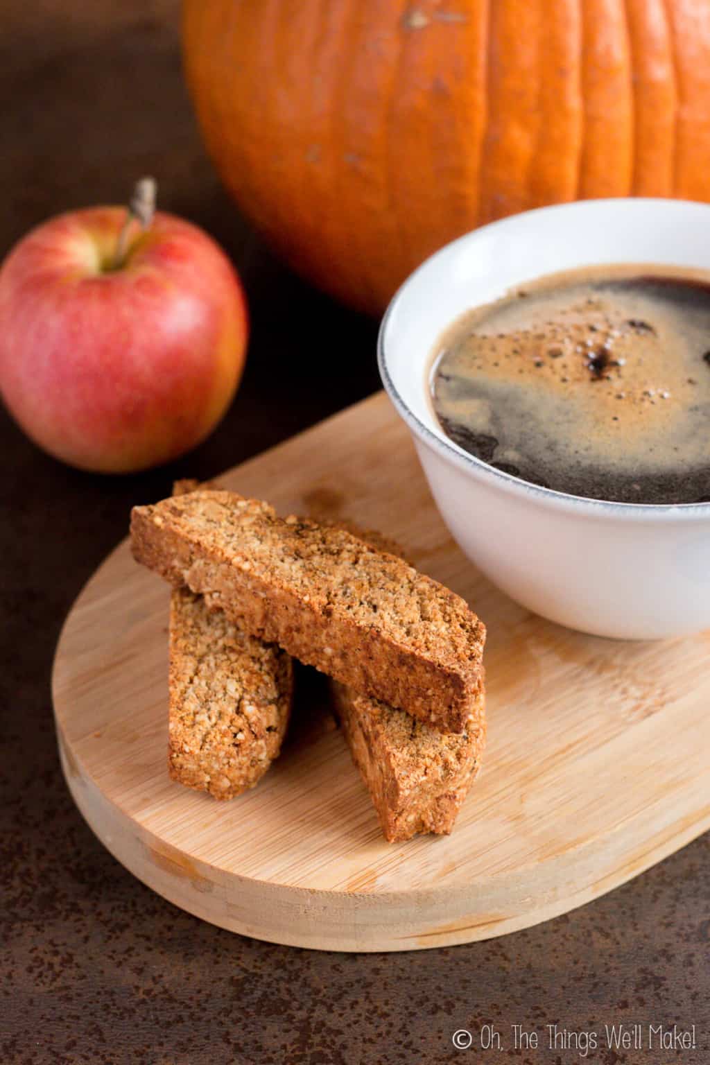 Three pumpkin spice biscotti on a wooden board with a cup of coffee on the side, and an apple in the background.