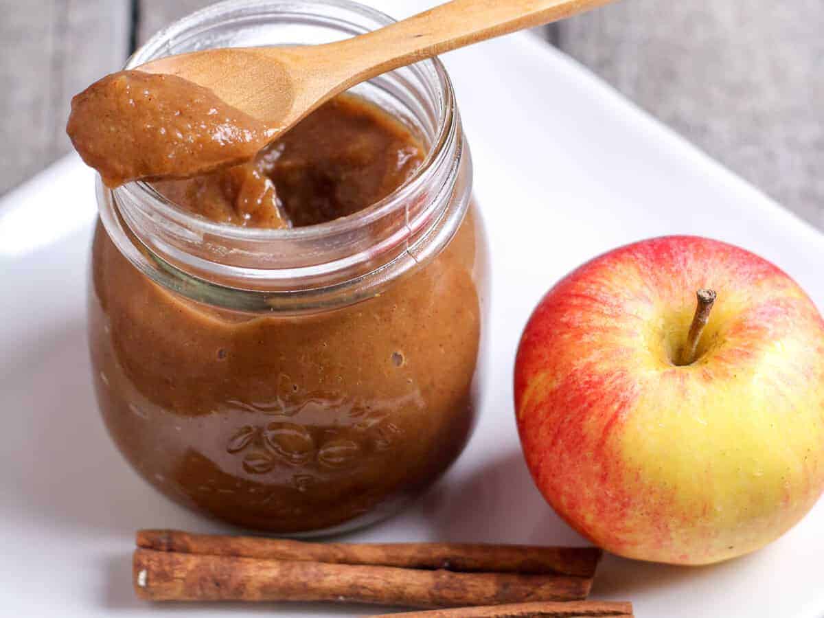 A spoonful of brown apple butter placed on top of a clear jar filled more apple butter placed next to a red-yellow apple and two cinnamon sticks. All of these placed on a white square plate.