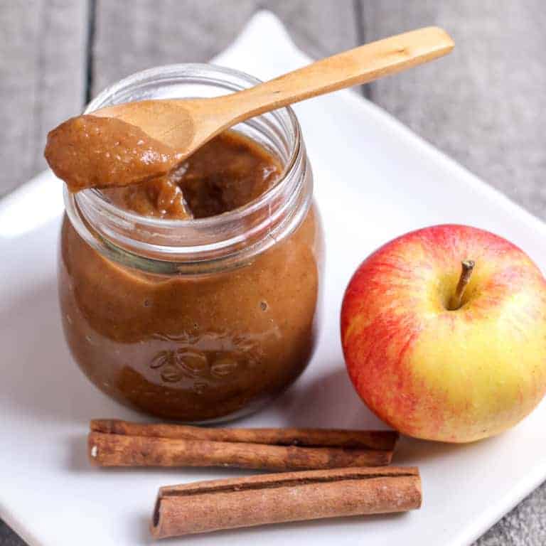 A spoonful of brown apple butter placed on top of a clear jar filled more apple butter placed next to a red-yellow apple and two cinnamon sticks. All of these placed on a white square plate.