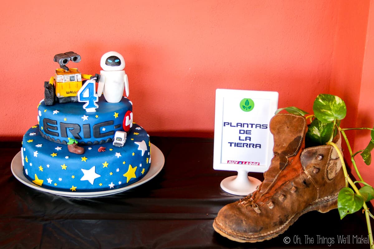 A table with Wall-E themed cake and decor. On the left is a blue fondant cake decorated with white and yellow stars, Wall-E and Eve fondant decor on top, and a number four candle. On the right is a brown boot with a plant vine tucked in it and a picture frame beside it saying "plantas de la tierra."