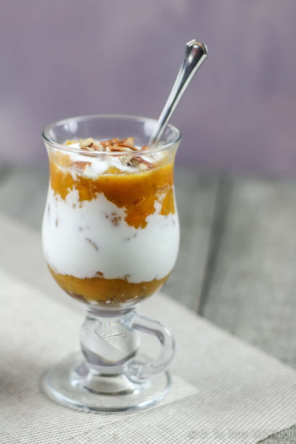 A glass full of pumpkin layered dessert of pumpkin puree and yogurt, topped off with chopped pecans and a silver spoon in it leaning to the side.