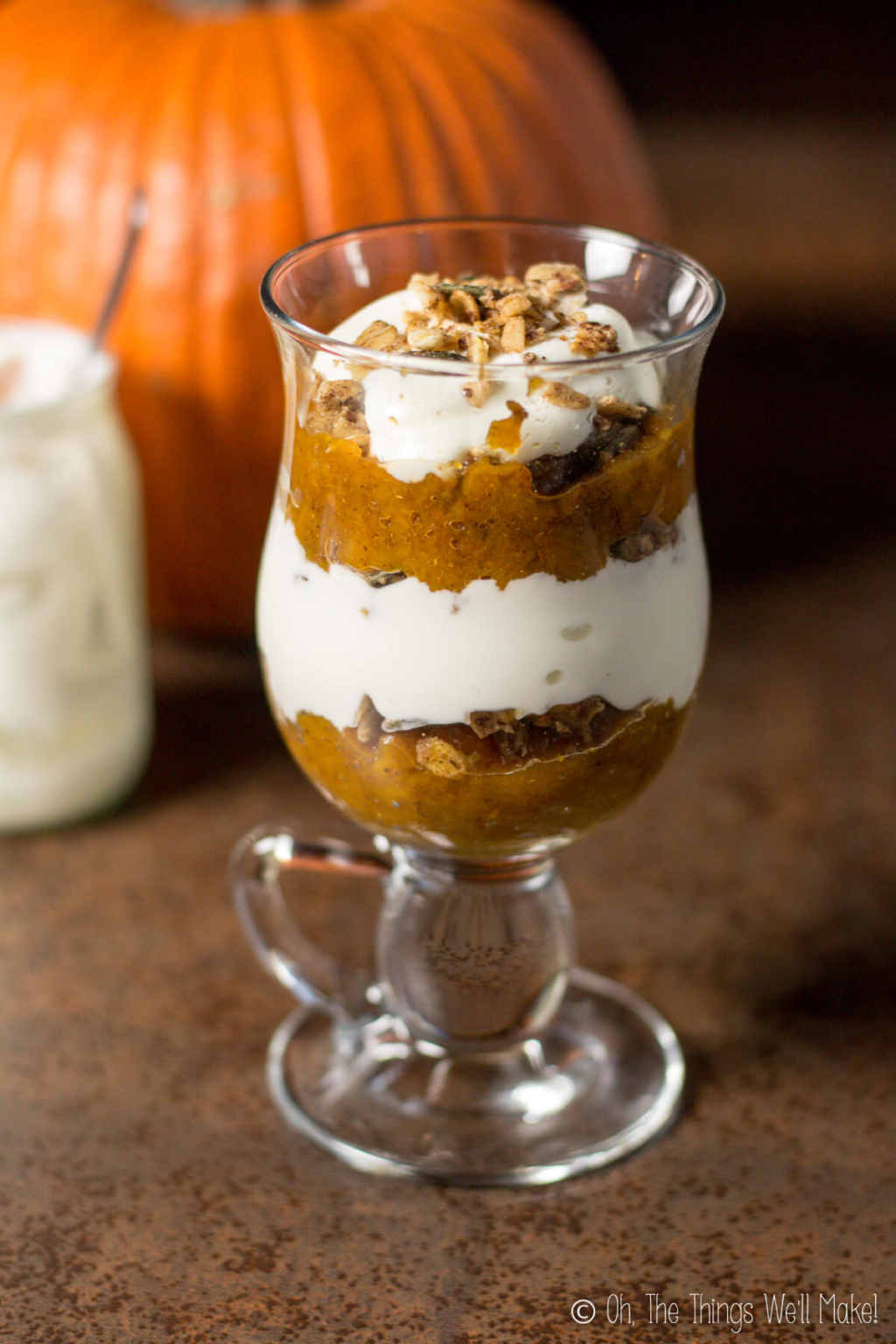 A glass full of pumpkin layered dessert of pumpkin puree and yogurt, topped off with chopped pecans. This glass is place in front of an orange pumpkin and a yogurt container.
