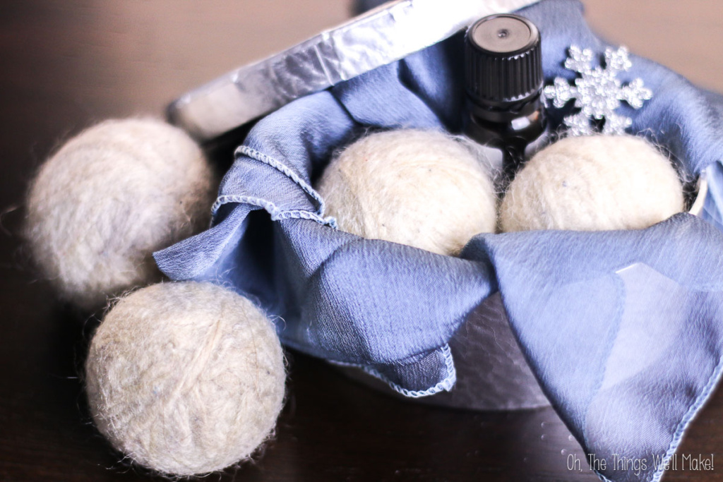 A small tin with a blue cloth holds diy wool dryer balls, a bottle of essential oil, and a decorative silver snowflake on a dark surface.