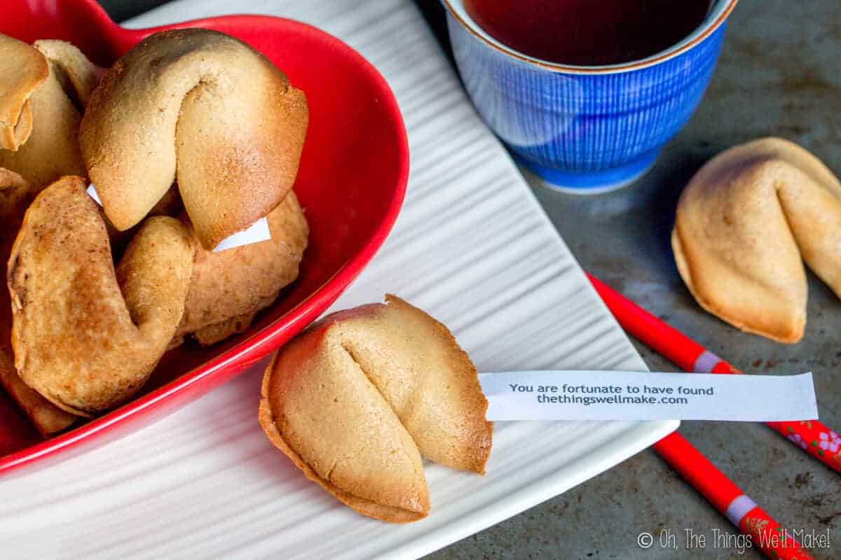 Overhead view of several homemade fortune cookies in a heart-shaped red bowl next to a cup of tea and a separate fortune cookie. One has a fortune that says "You are fortunate to have found thethingswellmake.com."