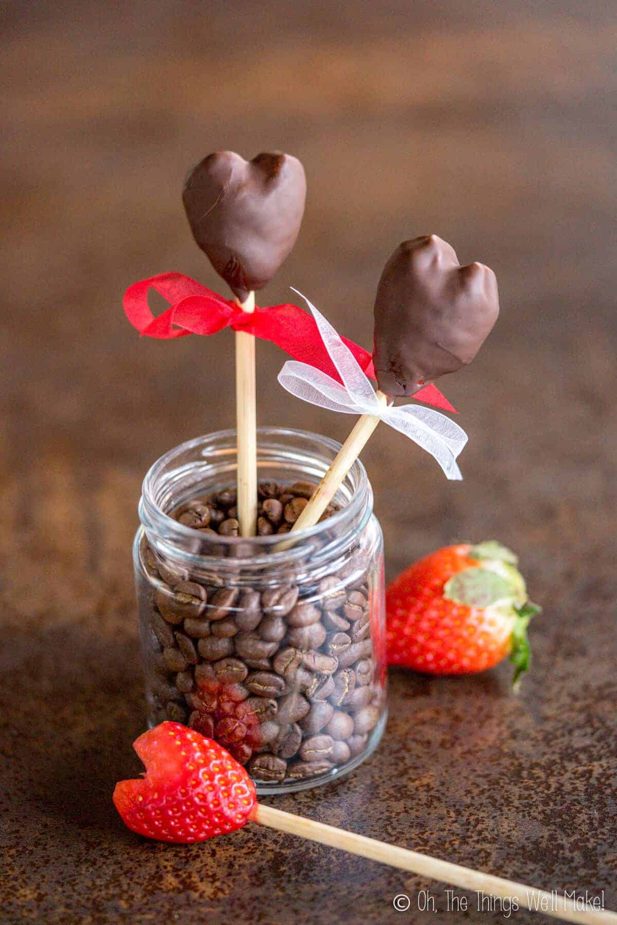 Two chocolate covered strawberries in a heart shape and next to a heart shaped strawberry on a stick ready to be dipped in chocolate.