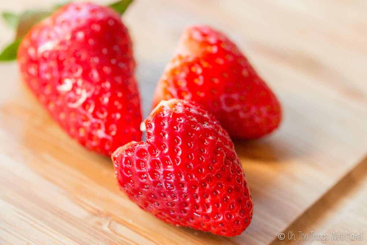 3 strawberreis on a bamboo cutting board, two of them have been cut into heart shapes