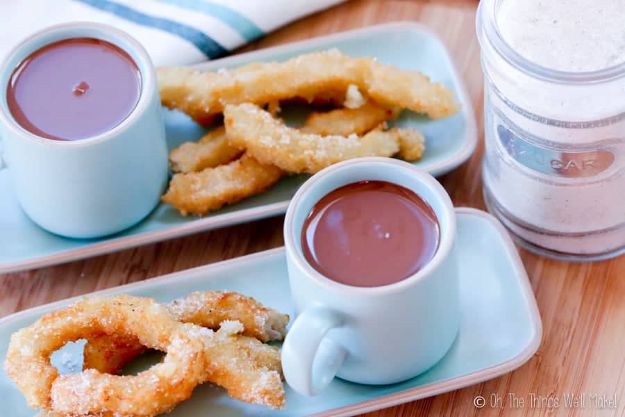 Overhead view of two plates of churros sprinkled with sugar with cups filled with Spanish hot chocolate.