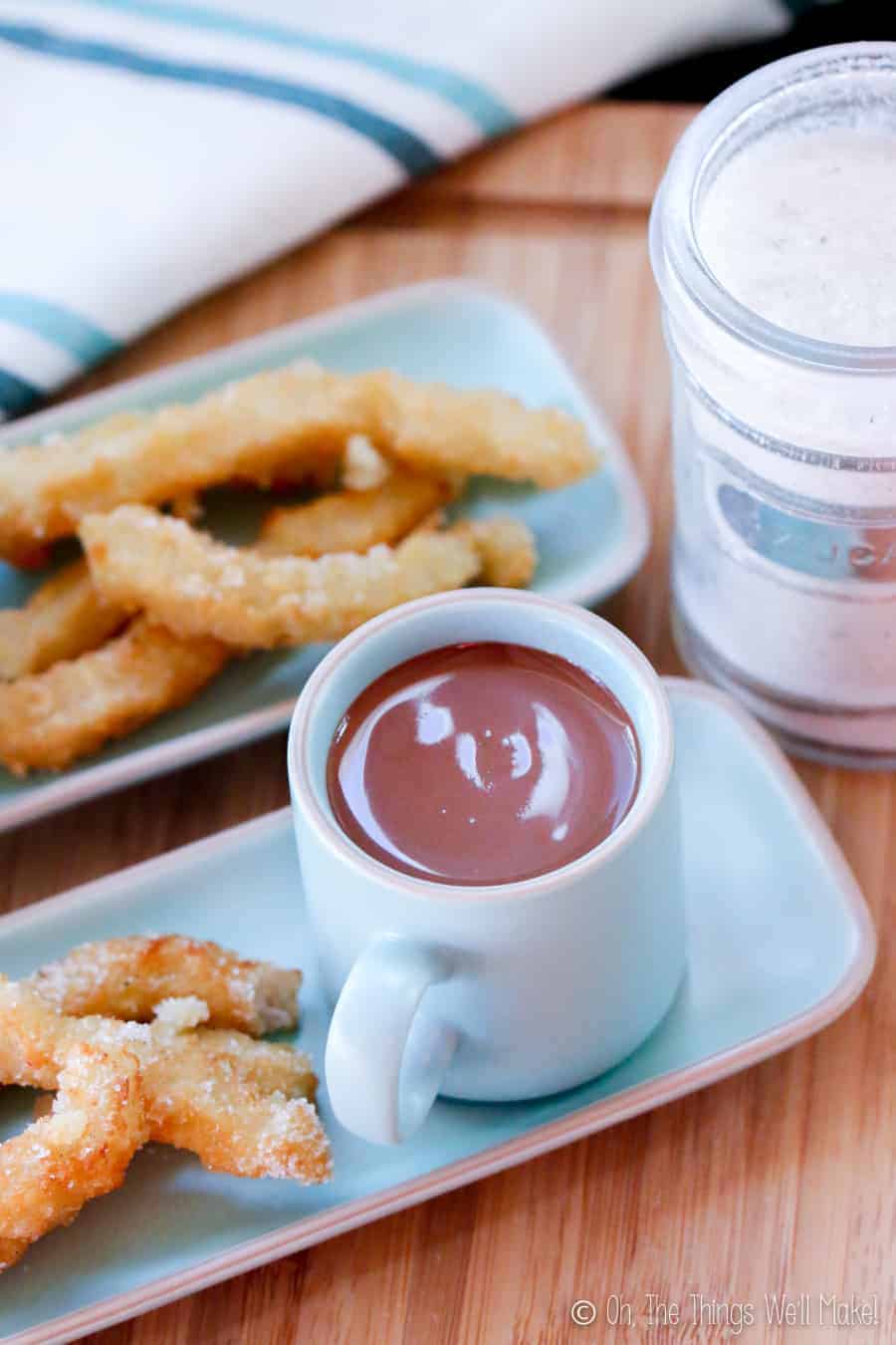 overhead view of 2 plates of churros with chocolate and a jar filled with sugar.