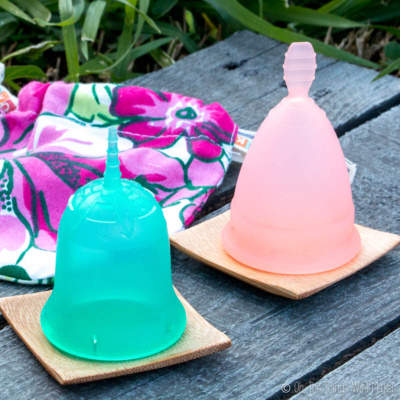 Two menstrual cups, one green Sckoon and one pink generic placed on square wooden platters with a floral bag behind them.