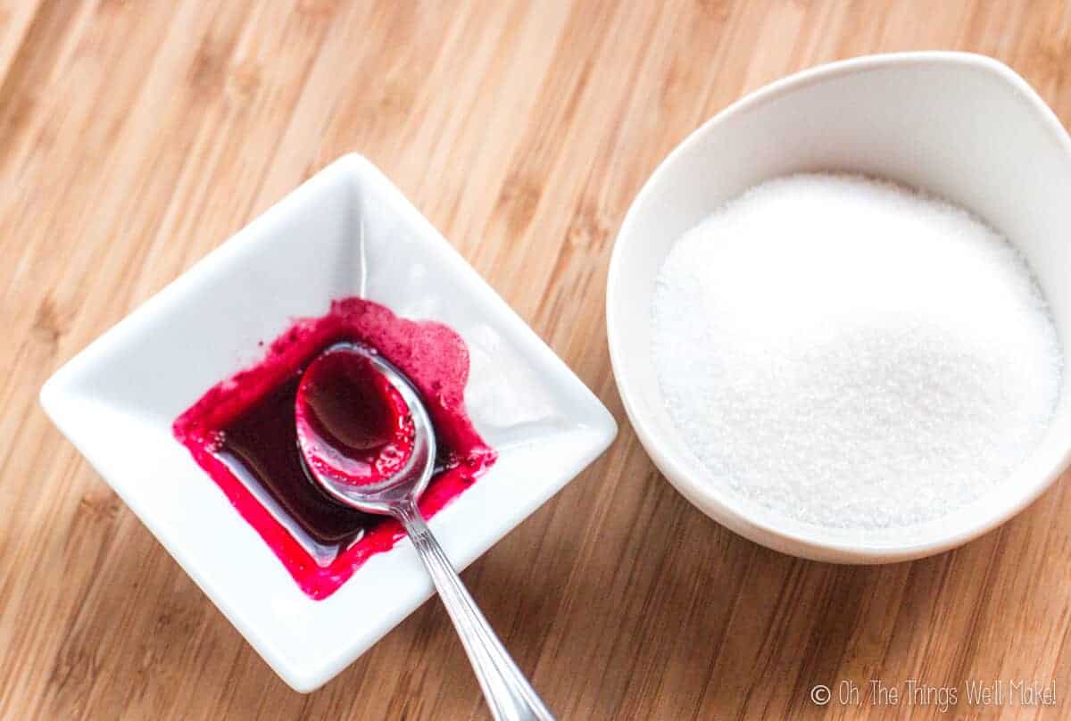 A small bowl of pink coloring on left next to a bowl of white sugar on the right.