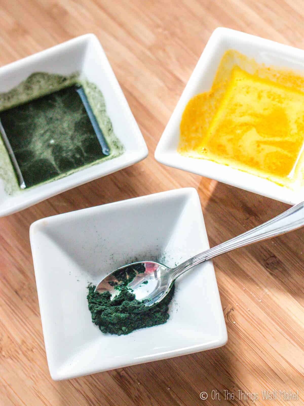 Overhead view of a small bowl with spirulina in front of 2 bowls, one with yellow coloring and another with green colorant