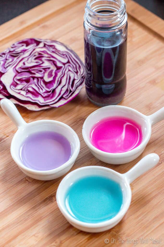 3 bowls of liquid, one turquoise, one pink, and one purple, in front of a bottle of liquid made from red cabbage, and next to a fresh red cabbage head sliced in half.