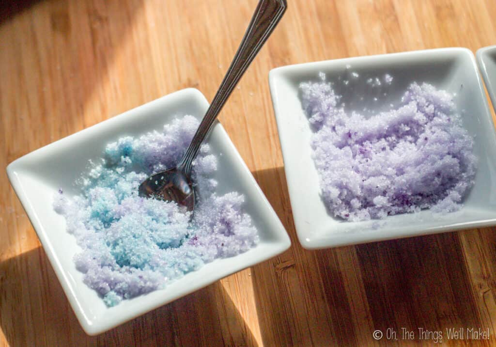 2 bowls of colored sugar crystals. The one on the right has purple sugar crystals. The one on the right is a bowl of purple sugar crystals with a spoon and the crystals around the spoon are light blue.