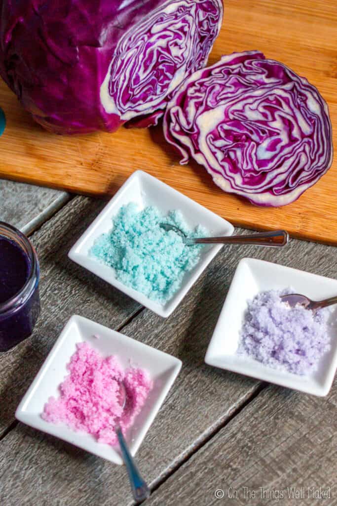 3 small bowls of colored sugar crystals, one pink, one purple, and one light blue, in front of a sliced open red cabbage on a bamboo cutting board.