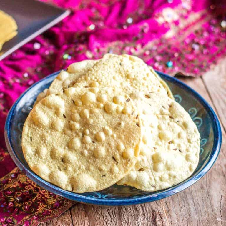 Close-up of a bowl full of homemade papadums made from whole urids. A bright pink fabric is place on the background of the bowl.