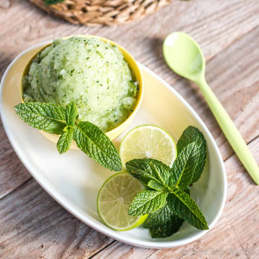 Overhead view of a mojito sorbet in a yellow bowl, garnished with spearmint and lime halves.