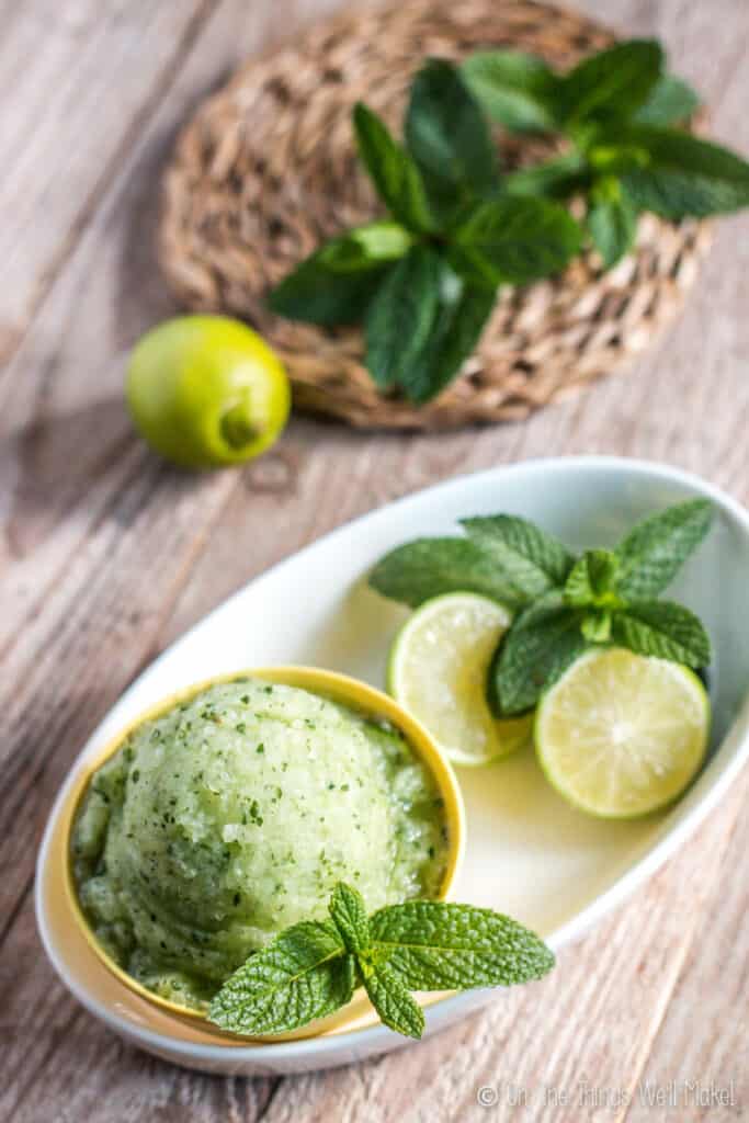 Overhead view of a mojito sorbet in a yellow bowl, garnished with spearmint and lime halves.