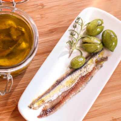 Overhead view of a plate of homemade salt cured anchovy fillets with green olives on the side, next to a jar filled with anchovies covered in virgin olive oil and salt mixture.