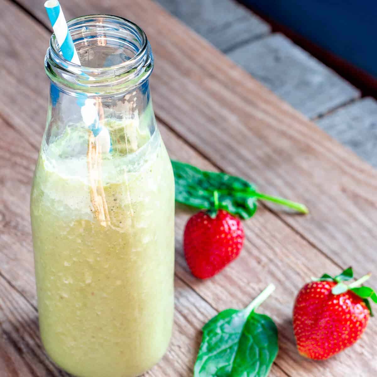 A bottle full of spinach smoothie with a blue and white straw inside, and a couple of strawberries scattered around the bottle.