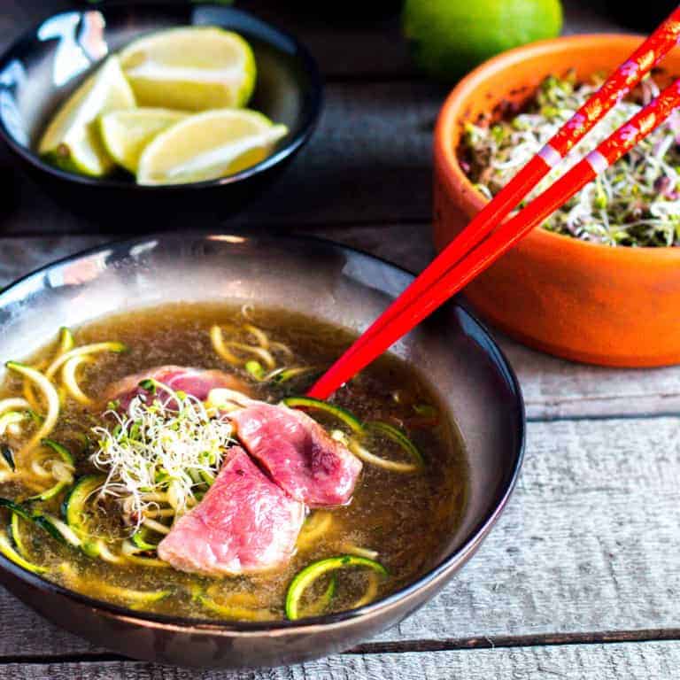 A black bowl full of homemade Vietnamese pho made of beef broth, zucchini noodles, and slices of beef steak, with red chopsticks on the side. Bowls of lime and garnish are in the background.