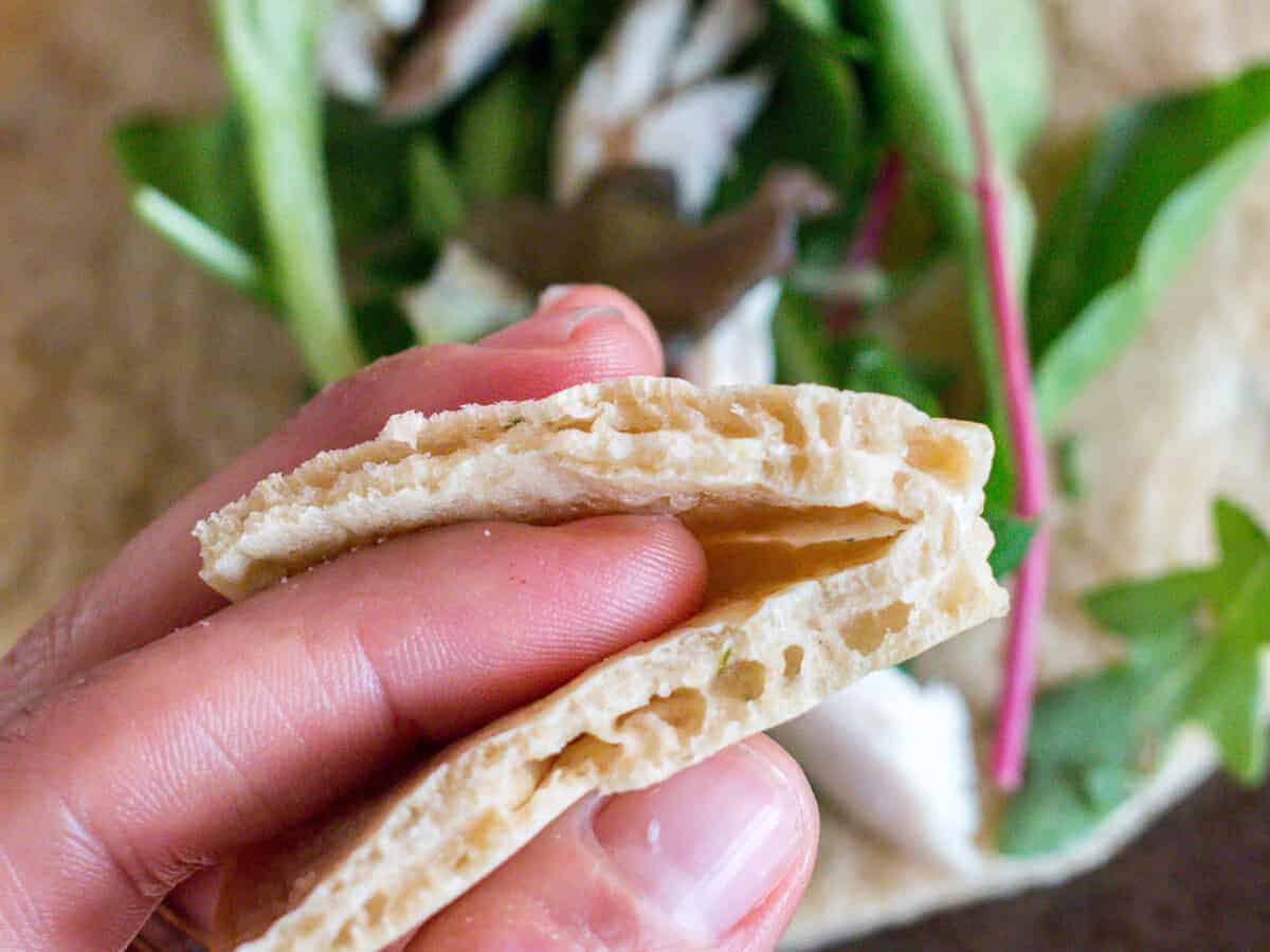 Close-up overhead view of a hand holding a homemade paleo pita bread folded in half with a pita bread full of leafy greens in the background.
