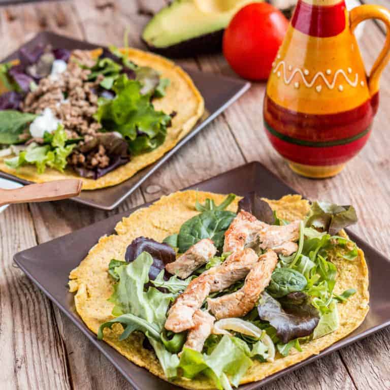 Two plates full of garnished paleo tortillas made with flaxseed and eggs. Foreground is a plate of tortillas topped with chicken and leafy greens. Background is a plate of tortillas topped with minced beef and leafy greens.