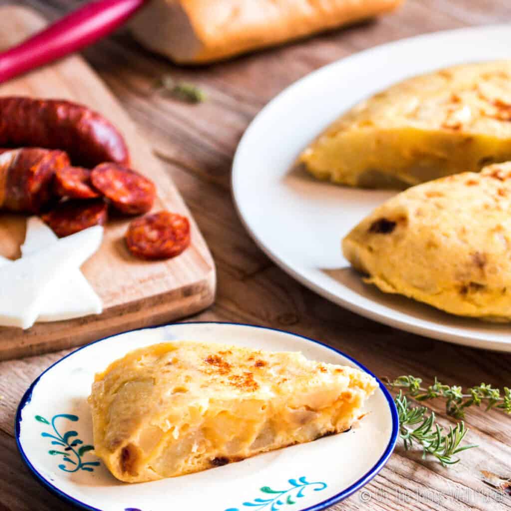 A slice of Spanish tortilla on a plate in front of some chorizo and cheese and the rest of the tortilla.