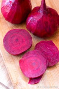 Beetrooot slices on a bamboo cutting board in front of two peeled beets.